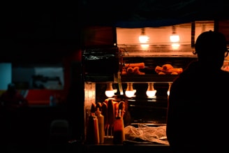 A dimly lit street food stall featuring churros under warm, glowing lights. The silhouette of a person is visible next to the stall, creating a contrast with the illuminated food items. Bottles of condiments are also present on the counter.
