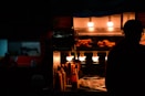 A dimly lit street food stall featuring churros under warm, glowing lights. The silhouette of a person is visible next to the stall, creating a contrast with the illuminated food items. Bottles of condiments are also present on the counter.