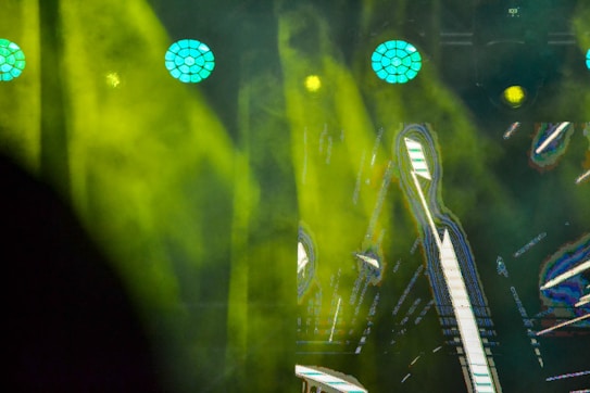 Bright yellow and green stage lights shine through smoke or fog, creating a vibrant and dynamic atmosphere. The background contains graphics with geometric shapes and lines, possibly on a screen.