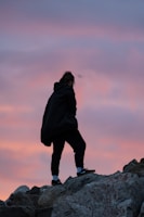 A person dressed in dark clothing is walking or climbing over rocky terrain. The sky behind them displays a beautiful blend of pink and purple hues, suggesting either dawn or dusk.