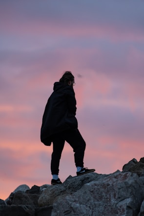 A person dressed in dark clothing is walking or climbing over rocky terrain. The sky behind them displays a beautiful blend of pink and purple hues, suggesting either dawn or dusk.