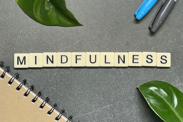 Tiles Spelling Out The Word 'Mindfulness' Are Arranged On A Gray Surface, Accompanied By A Few Green Leaves, A Notebook On The Lower Left Corner, And Two Pens To The Upper Right.