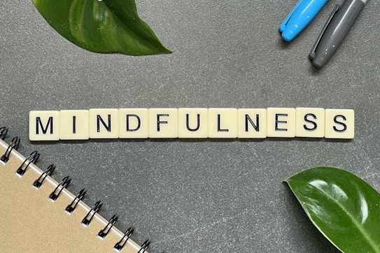 Tiles spelling out the word 'MINDFULNESS' are arranged on a gray surface, accompanied by a few green leaves, a notebook on the lower left corner, and two pens to the upper right.