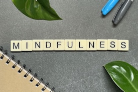 Tiles spelling out the word 'MINDFULNESS' are arranged on a gray surface, accompanied by a few green leaves, a notebook on the lower left corner, and two pens to the upper right.