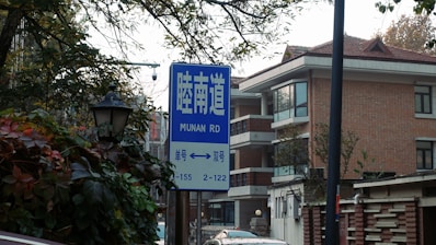 A street sign with text in both Chinese and English indicating 'Munan Rd' stands in front of a residential building with red brick walls and large windows. Surrounding the sign are trees with green and brown leaves, and there is a traditional street lamp surrounded by foliage.