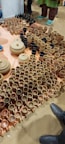 A variety of clay pots, mugs, and bowls are arranged on the floor. The ceramics are primarily in shades of brown and beige, with a few black items. Several people stand nearby, with only their shoes visible, indicating the setting is likely a market or exhibition.