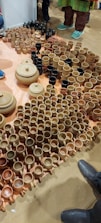 A variety of clay pots, mugs, and bowls are arranged on the floor. The ceramics are primarily in shades of brown and beige, with a few black items. Several people stand nearby, with only their shoes visible, indicating the setting is likely a market or exhibition.