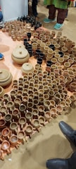 A variety of clay pots, mugs, and bowls are arranged on the floor. The ceramics are primarily in shades of brown and beige, with a few black items. Several people stand nearby, with only their shoes visible, indicating the setting is likely a market or exhibition.
