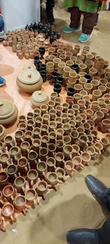 A variety of clay pots, mugs, and bowls are arranged on the floor. The ceramics are primarily in shades of brown and beige, with a few black items. Several people stand nearby, with only their shoes visible, indicating the setting is likely a market or exhibition.
