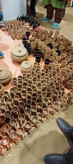 A variety of clay pots, mugs, and bowls are arranged on the floor. The ceramics are primarily in shades of brown and beige, with a few black items. Several people stand nearby, with only their shoes visible, indicating the setting is likely a market or exhibition.