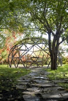 A wooden geodesic dome structure with seating is located in a garden, surrounded by lush greenery and trees. A stone path leads to the dome, and the area is shaded by tree branches with sunlight filtering through. Bright red and orange foliage is visible in the background.