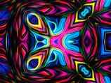 An intricate, symmetrical pattern that resembles a kaleidoscope. Vibrant colors including blues, pinks, and yellows swirl and blend together, creating a dynamic and psychedelic visual effect. The design features layered geometric shapes and curves, giving a sense of depth and movement.