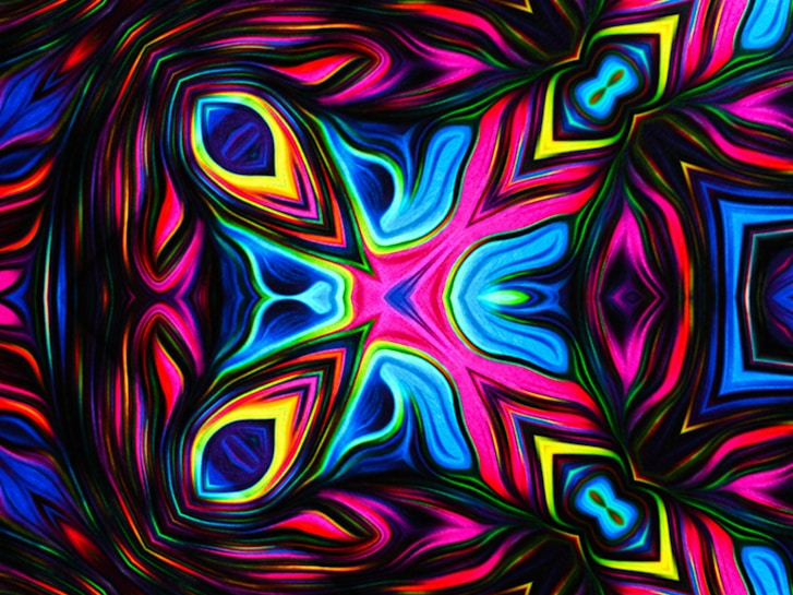 An intricate, symmetrical pattern that resembles a kaleidoscope. Vibrant colors including blues, pinks, and yellows swirl and blend together, creating a dynamic and psychedelic visual effect. The design features layered geometric shapes and curves, giving a sense of depth and movement.
