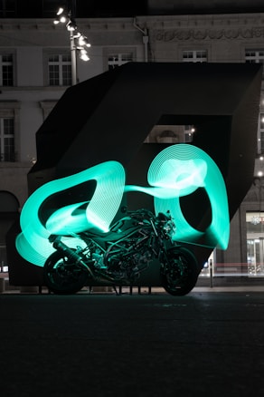 A motorcycle is positioned in front of a large, dark, abstract sculpture. Bright green light trails swirl around the motorcycle, creating dynamic patterns that contrast with the surrounding dark setting. The background features a building with illuminated windows and architectural details.