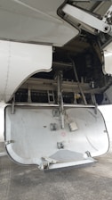 The image depicts the open undercarriage panel of an aircraft, with visible mechanical components and interior structure. The panel shows signs of wear and exposure to dirt, suggesting usage. The background includes parts of the aircraft's wing and various hydraulic and structural elements.