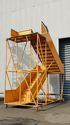 A mobile staircase structure with a metal frame and wheels, painted in bright yellow, stands against a corrugated metal building.