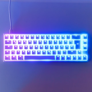A compact mechanical keyboard with RGB lighting illuminating the keys. The colors transition from pink on the left side to blue on the right side, creating a vibrant and dynamic visual effect. The keyboard layout includes a QWERTZ configuration, common in German-speaking countries.