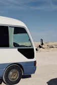 A partially visible white and blue minibus is parked on a desert-like terrain with a clear blue sky above. The bus has a side window with a curtain and a black mirror extending from the door.