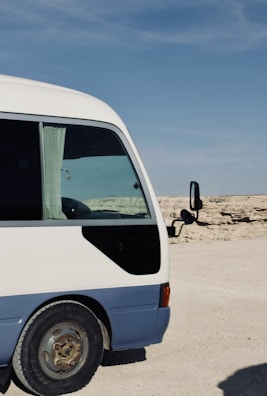 A partially visible white and blue minibus is parked on a desert-like terrain with a clear blue sky above. The bus has a side window with a curtain and a black mirror extending from the door.