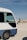 A partially visible white and blue minibus is parked on a desert-like terrain with a clear blue sky above. The bus has a side window with a curtain and a black mirror extending from the door.