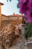 A family stands together in a rustic village setting with stone walls and houses. The scene is framed by vibrant purple flowers, adding a splash of color. The family consists of two adults and a child, all dressed in light, casual clothing, and they appear happy and relaxed.