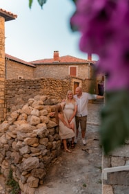 A family stands together in a rustic village setting with stone walls and houses. The scene is framed by vibrant purple flowers, adding a splash of color. The family consists of two adults and a child, all dressed in light, casual clothing, and they appear happy and relaxed.