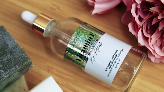 A glass dropper bottle with a gold cap, featuring a label that includes the words 'Vitamin E' and 'Facial Serum.' The bottle is lying on a wooden surface next to some pink flowers and a green bar of soap.