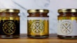 Three jars of natural sweet honey are displayed on a wooden surface. Each jar has a gold lid and is labeled with a bee-themed design. The left jar has a black label, the middle jar has a white label, and the right jar also features a white label with gold accents. The labels read 'Organic Pure Product' with images of honey dippers and a bee.