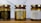 Three jars of natural sweet honey are displayed on a wooden surface. Each jar has a gold lid and is labeled with a bee-themed design. The left jar has a black label, the middle jar has a white label, and the right jar also features a white label with gold accents. The labels read 'Organic Pure Product' with images of honey dippers and a bee.