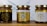 Three jars of natural sweet honey are displayed on a wooden surface. Each jar has a gold lid and is labeled with a bee-themed design. The left jar has a black label, the middle jar has a white label, and the right jar also features a white label with gold accents. The labels read 'Organic Pure Product' with images of honey dippers and a bee.