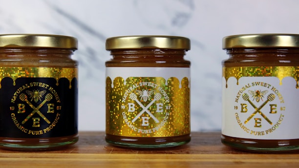 Three jars of natural sweet honey are displayed on a wooden surface. Each jar has a gold lid and is labeled with a bee-themed design. The left jar has a black label, the middle jar has a white label, and the right jar also features a white label with gold accents. The labels read 'Organic Pure Product' with images of honey dippers and a bee.