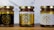 Three jars of natural sweet honey are displayed on a wooden surface. Each jar has a gold lid and is labeled with a bee-themed design. The left jar has a black label, the middle jar has a white label, and the right jar also features a white label with gold accents. The labels read 'Organic Pure Product' with images of honey dippers and a bee.