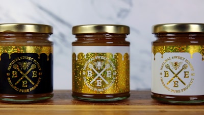 Three jars of natural sweet honey are displayed on a wooden surface. Each jar has a gold lid and is labeled with a bee-themed design. The left jar has a black label, the middle jar has a white label, and the right jar also features a white label with gold accents. The labels read 'Organic Pure Product' with images of honey dippers and a bee.