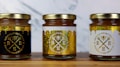 Three jars of natural sweet honey are displayed on a wooden surface. Each jar has a gold lid and is labeled with a bee-themed design. The left jar has a black label, the middle jar has a white label, and the right jar also features a white label with gold accents. The labels read 'Organic Pure Product' with images of honey dippers and a bee.