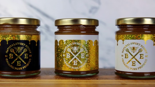 Three jars of natural sweet honey are displayed on a wooden surface. Each jar has a gold lid and is labeled with a bee-themed design. The left jar has a black label, the middle jar has a white label, and the right jar also features a white label with gold accents. The labels read 'Organic Pure Product' with images of honey dippers and a bee.
