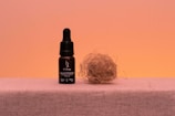A bottle with a dropper labeled 'H Drop' sits on a flat surface next to a bundle of dried twigs or straw, against a warm peach-colored background.