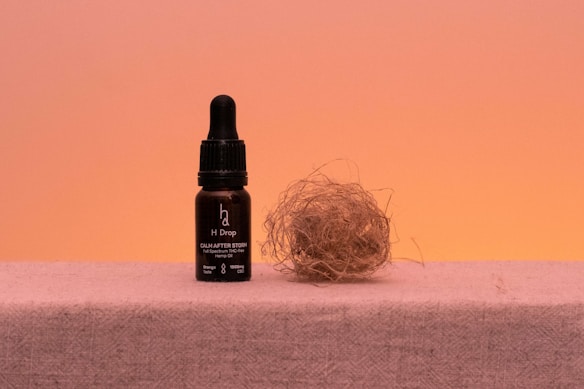 A bottle with a dropper labeled 'H Drop' sits on a flat surface next to a bundle of dried twigs or straw, against a warm peach-colored background.