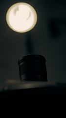 A camera lens is positioned prominently in the foreground with a soft-focus, bright light source above it. The background is dark and blurred, creating a dramatic contrast between the illuminated lens and its surroundings.