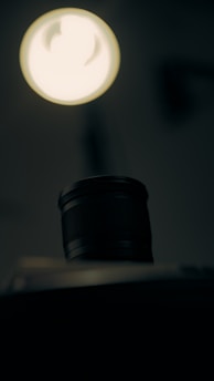 A camera lens is positioned prominently in the foreground with a soft-focus, bright light source above it. The background is dark and blurred, creating a dramatic contrast between the illuminated lens and its surroundings.