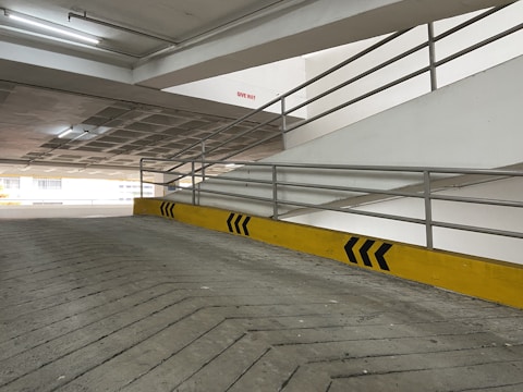 A concrete ramp in a parking garage with yellow and black directional arrows painted on a barrier. Metal railings line the sides of the ramp, and fluorescent lighting is visible on the ceiling. A red 'Give Way' sign is mounted on a wall.
