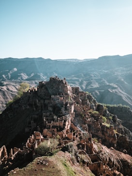 A sprawling ancient village built on a rugged hillside with numerous stone structures that appear to be ruins. The landscape extends into rolling hills and distant mountains under a clear blue sky, creating a sense of isolation and historical intrigue.
