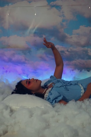 A person lies on a bed surrounded by fluffy clouds with an arm raised gently into the air. The individual is dressed in a light blue outfit with lace details and wears visible makeup with a focus on pink hues. The background is a sky filled with clouds, creating a dreamy and surreal atmosphere.