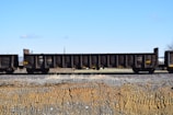 A train car with open-topped brown cargo wagons resting on railway tracks, set against a clear blue sky and a barren landscape with brown and gray gravel.