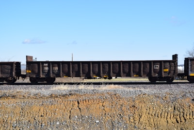 A train car with open-topped brown cargo wagons resting on railway tracks, set against a clear blue sky and a barren landscape with brown and gray gravel.