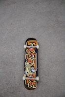 A skateboard lies on a rough, gray textured ground surface. The skateboard deck features a bold design with the word 'CREATURE' in large, stylized letters in orange and yellow hues. The center of the deck displays an illustration of a character or creature in a dynamic pose.