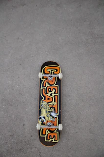 A skateboard lies on a rough, gray textured ground surface. The skateboard deck features a bold design with the word 'CREATURE' in large, stylized letters in orange and yellow hues. The center of the deck displays an illustration of a character or creature in a dynamic pose.