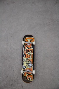A skateboard lies on a rough, gray textured ground surface. The skateboard deck features a bold design with the word 'CREATURE' in large, stylized letters in orange and yellow hues. The center of the deck displays an illustration of a character or creature in a dynamic pose.