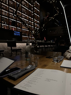 A dimly lit indoor scene of a restaurant or bar with a wooden table in the foreground displaying menus, glasses, and a napkin with utensils. In the background, a well-organized wall filled with bottles, illuminated by soft lighting, adds a cozy ambiance. A decorative element resembling a tree with string lights is visible, contributing to the atmosphere.