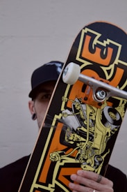 A person holding a skateboard vertically. The skateboard features a dynamic graphic of a car with a skeleton driving it, adorned with bold colors and a cartoonish style. The person is partially obscured by the skateboard, wearing a black cap and ear gauges. The background is a plain, off-white wall.