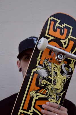 A person holding a skateboard vertically. The skateboard features a dynamic graphic of a car with a skeleton driving it, adorned with bold colors and a cartoonish style. The person is partially obscured by the skateboard, wearing a black cap and ear gauges. The background is a plain, off-white wall.